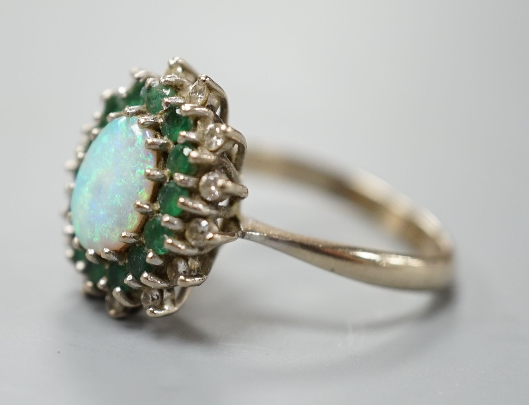 An 18ct white metal, white opal, emerald and diamond set oval cluster ring, size O, gross weight 6.2 grams.